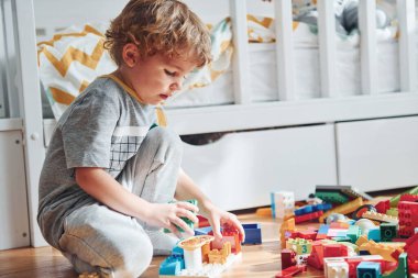Cute little boy sitting and have fun indoors in the bedroom with plastic construction set.
