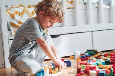 Cute little boy sitting and have fun indoors in the bedroom with plastic construction set.