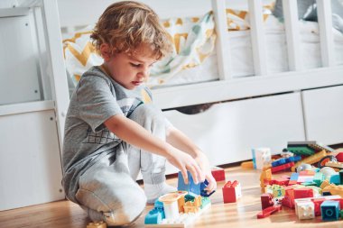 Cute little boy sitting and have fun indoors in the bedroom with plastic construction set.