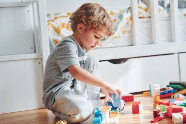 Cute little boy sitting and have fun indoors in the bedroom with plastic construction set.