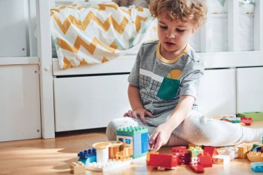 Cute little boy sitting and have fun indoors in the bedroom with plastic construction set.