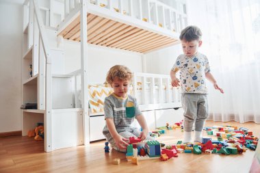 Two little boys have fun indoors in the bedroom with plastic construction set.