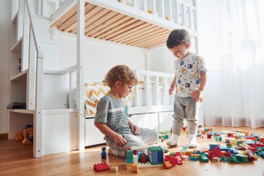 Two little boys have fun indoors in the bedroom with plastic construction set.