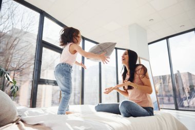Having fun with pillow on bed. Young mother with her daughter spending weekend together in room.