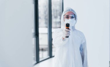 Kızılötesi termometre tutuyor. Laboratuvar önlüklü, koruyucu gözlüklü ve maskeli kadın doktor portresi..