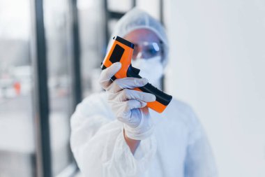Kızılötesi termometre tutuyor. Laboratuvar önlüklü, koruyucu gözlüklü ve maskeli kadın doktor portresi..