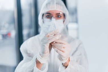 Şırınga tutuyor. Laboratuvar önlüklü, koruyucu gözlüklü ve maskeli kadın doktor portresi..