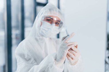 Holds syringe. Portrait of female doctor scientist in lab coat, defensive eyewear and mask.