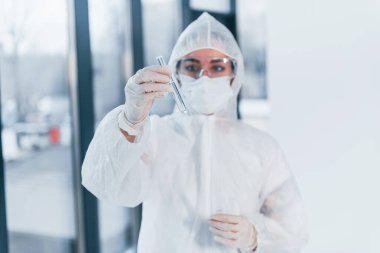 Holds test tube. Portrait of female doctor scientist in lab coat, defensive eyewear and mask.