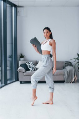 Ayakta duruyor ve minderi tutuyor. Spor kıyafetleri içinde ince vücutlu genç bir kadın evde spor yapıyor..
