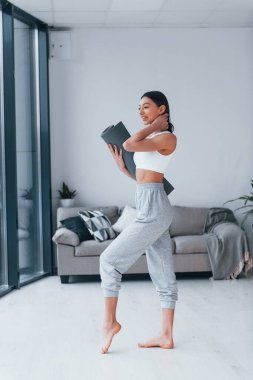 Ayakta duruyor ve minderi tutuyor. Spor kıyafetleri içinde ince vücutlu genç bir kadın evde spor yapıyor..