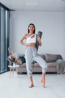 Ayakta duruyor ve minderi tutuyor. Spor kıyafetleri içinde ince vücutlu genç bir kadın evde spor yapıyor..