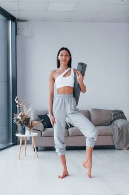 Ayakta duruyor ve minderi tutuyor. Spor kıyafetleri içinde ince vücutlu genç bir kadın evde spor yapıyor..