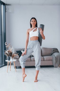 Ayakta duruyor ve minderi tutuyor. Spor kıyafetleri içinde ince vücutlu genç bir kadın evde spor yapıyor..
