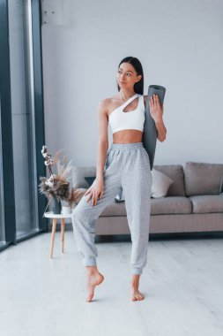 Ayakta duruyor ve minderi tutuyor. Spor kıyafetleri içinde ince vücutlu genç bir kadın evde spor yapıyor..