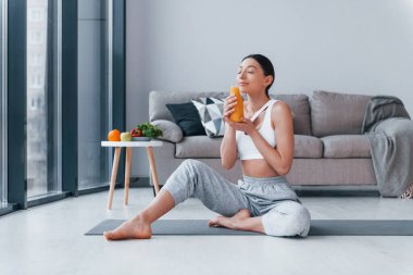 Elinde portakal suyu olan bir bardak tutuyor. Spor kıyafetleri içinde ince vücutlu genç bir kadın evde spor yapıyor..
