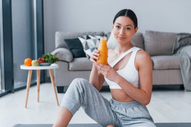 Elinde portakal suyu olan bir bardak tutuyor. Spor kıyafetleri içinde ince vücutlu genç bir kadın evde spor yapıyor..