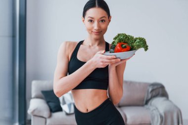 Sağlıklı yiyecekleri ellerinde tutuyor. Siyah spor giyimli zayıf vücutlu genç bir kadın evde spor salonuna gidiyor..
