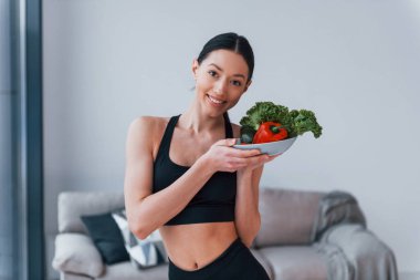 Sağlıklı yiyecekleri ellerinde tutuyor. Siyah spor giyimli zayıf vücutlu genç bir kadın evde spor salonuna gidiyor..