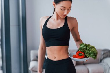 Sağlıklı yiyecekleri ellerinde tutuyor. Siyah spor giyimli zayıf vücutlu genç bir kadın evde spor salonuna gidiyor..