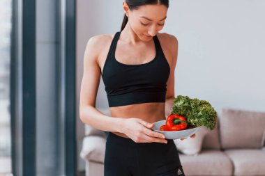 Sağlıklı yiyecekleri ellerinde tutuyor. Siyah spor giyimli zayıf vücutlu genç bir kadın evde spor salonuna gidiyor..