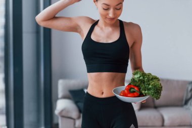 Sağlıklı yiyecekleri ellerinde tutuyor. Siyah spor giyimli zayıf vücutlu genç bir kadın evde spor salonuna gidiyor..