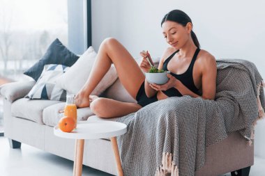 Young woman with slim body shape in sportswear sits on sofa and eats healthy diet food indoors at home.
