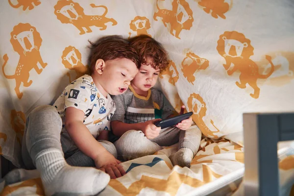 İki küçük çocuk dinleniyor ve akıllı telefonlarıyla birlikte battaniyenin altında eğleniyorlar..