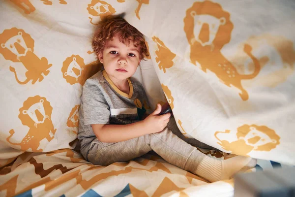 かわいい男の子が休んで 手に携帯電話で毛布の下で屋内で楽しみを持っています — ストック写真