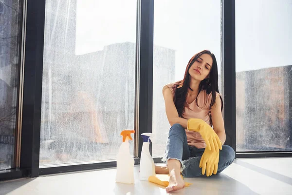 Woman sits on window sill with bottles of cleaning spray and with gloves.