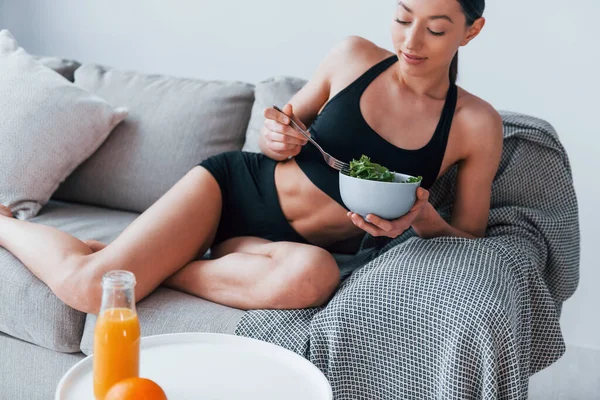 운동복 차림의 몸매의 여자는 소파에 집안에서 건강에 음식을 먹는다 — 스톡 사진