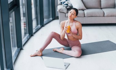Spor kıyafetleri içinde ince vücutlu genç bir kadın evde portakal suyu ve dizüstü bilgisayarla mindere oturuyor..