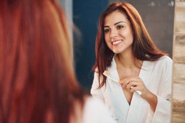 Güzel genç bir kadın banyoda aynanın yanında duruyor ve dişlerini fırçalıyor..