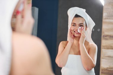 Beautiful young woman standing in bathroom, looking into the mirror and taking care of her face.