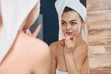 Beautiful young woman standing in bathroom, looking into the mirror and taking care of her face.