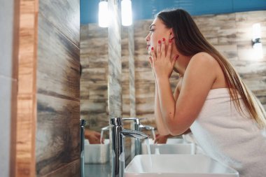 Beautiful young woman standing in bathroom near the mirror and washing her face.