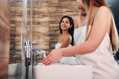 Beautiful young woman standing in bathroom near the mirror and washing hands. Friends behind.