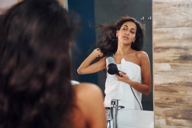 Güzel genç bir kadın banyoda aynanın yanında duruyor ve saçlarını kurutuyor..