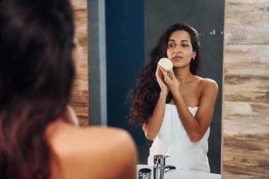 Güzel esmer kadın banyoda aynanın yanında duruyor ve yüzünü temizliyor..