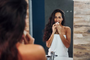 Güzel esmer kadın banyoda aynanın yanında duruyor ve yüzünü temizliyor..