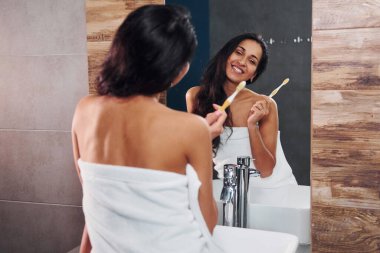 Beautiful young brunette standing in bathroom near the mirror and brushing her teeth.