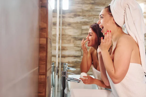 两个漂亮的年轻女子站在镜子旁边的浴室里 擦拭着她们的脸 — 图库照片