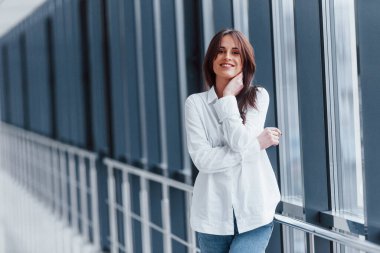 Gündüz vakti modern havaalanında ya da koridorda beyaz gömlekli esmer kadın parmaklıklara yaslanıyor..