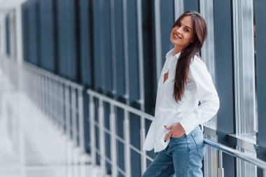 Gündüz vakti modern havaalanında ya da koridorda beyaz gömlekli esmer kadın parmaklıklara yaslanıyor..