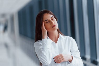 Brunette in white shirt standing indoors in modern airport or hallway at daytime and having headache.