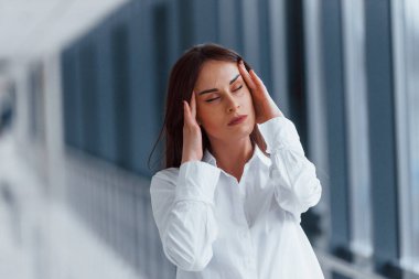 Brunette in white shirt standing indoors in modern airport or hallway at daytime and having headache.