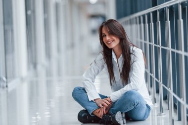 Brunette in white shirt sits indoors in modern airport or hallway at daytime.