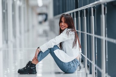 Beyaz gömlekli esmer, modern havaalanında ya da gündüz vakti koridorda oturuyor..