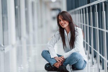 Beyaz gömlekli esmer, modern havaalanında ya da gündüz vakti koridorda oturuyor..