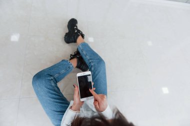 Gündüz vakti modern havaalanında ya da koridorda oturan ellerinde telefon olan beyaz tişörtlü esmer kız manzarası..